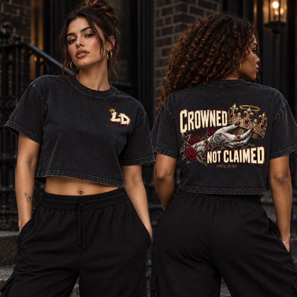 Two women modeling the washed-black Lions Den Crowned Not Claimed mineral-wash crop tee, showing the crowned LD front mark and full back artwork