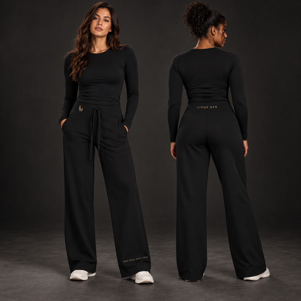 Two women model the black Lions Den MMXXVI straight-leg yoga pants, showing the LD hip mark, lower-leg motto, and back waistband wordmark.